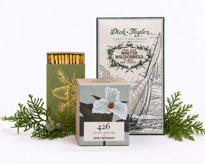 The Winter Glow: The Quiet Botanist 426 mini candle, a green box of matches with a gold bee crest, and Dick Taylor Winter Wilderness dark chocolate